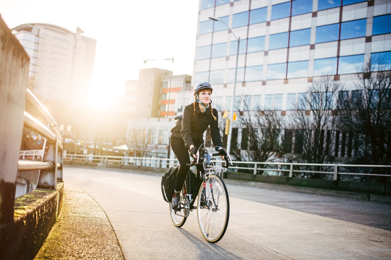 7 Easy Ways to Work Out on the Job as a Traveler A smiling young woman commuting in an urban city environment on her street bicycle, waterproof panniers on her bike rack. She rides up a ramp from downtown Portland, Oregon, that leads up to the Burnside bridge. Colorful sky from the setting sun. Horizontal image with copy space. A depiction of young adult / hipster lifestyle in the city.