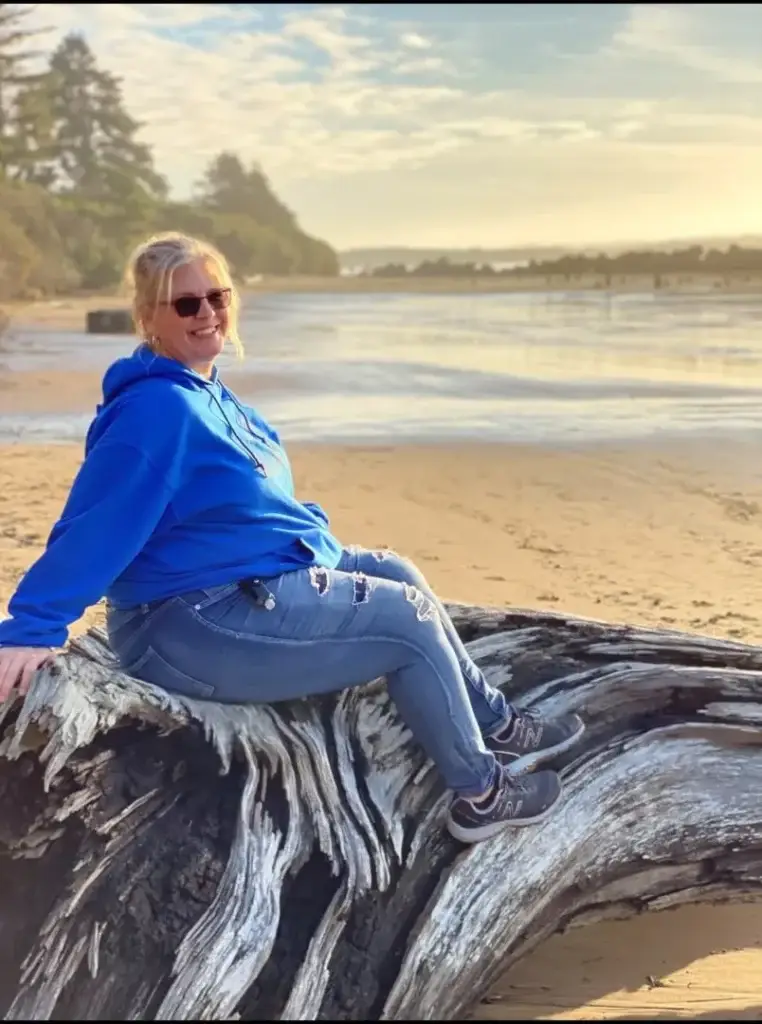 blonde woman sitting on fallen tree and taking photo with coast in the background