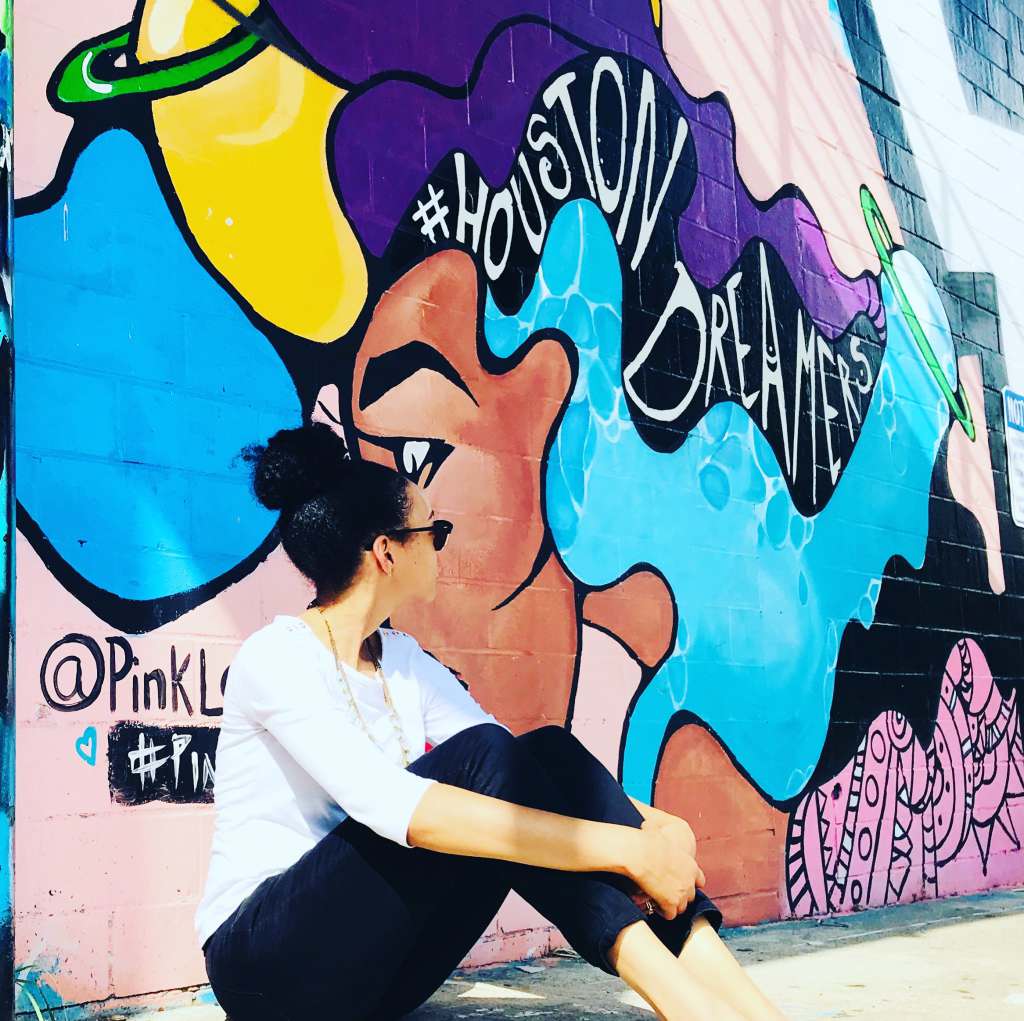 A woman sitting in front of a vibrant mural located on Leeland Street in Houston, Texas. The mural is created by an artist known as @PinkLomein. 